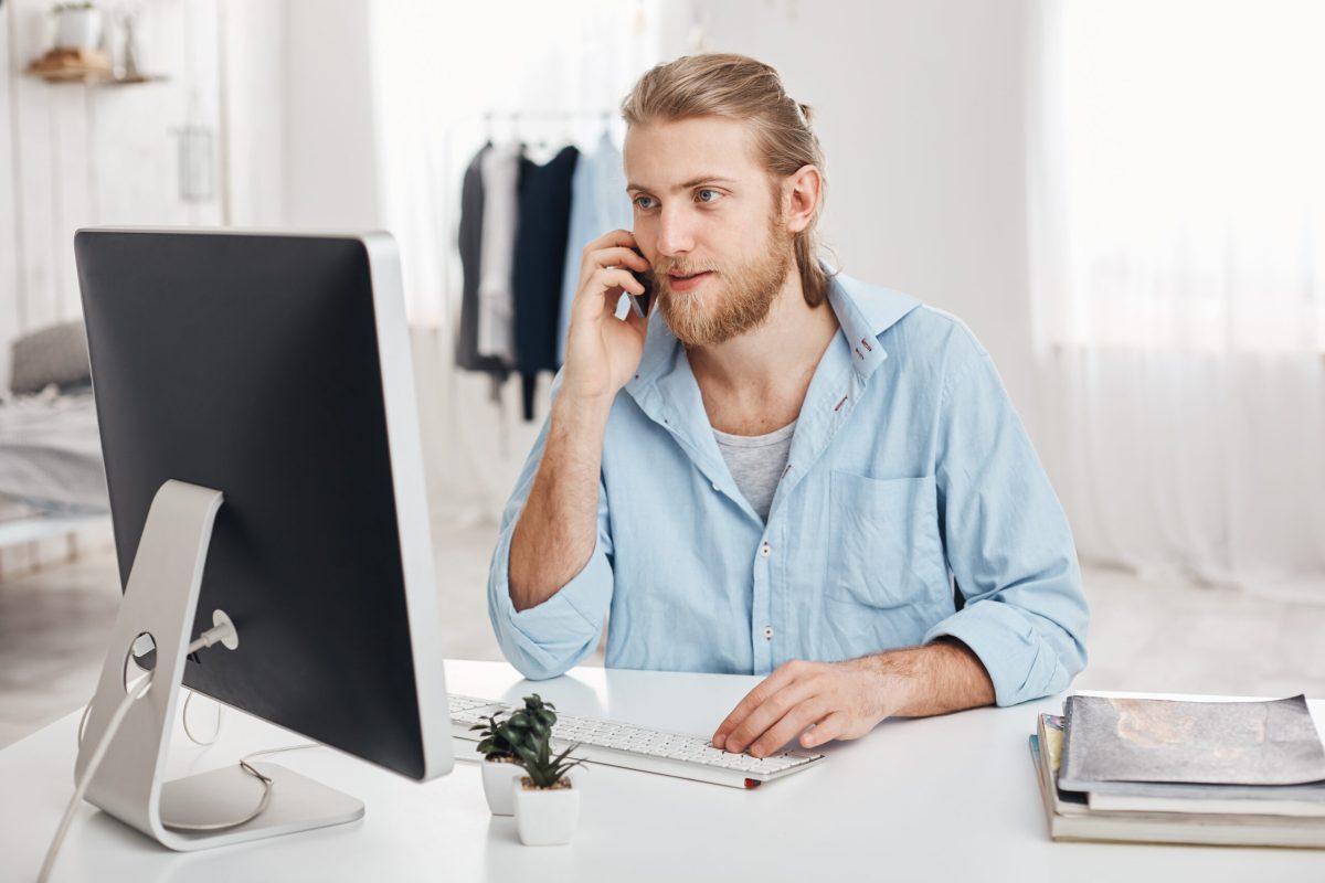 Bearded skilled young fair-haired businessman works on new project, sits in front of screen, has phone conversation, discusses last financial report with businesspartners. Office worker chats with boss